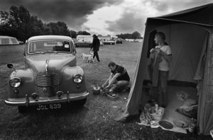 Two men campers prepare breakfast at the Sandy Balls campsite beside their vintage Austin A40 Devon car. A dog walker and storm clouds in the background. First published in The Sunday Times Date: 1989