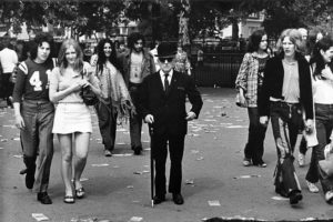 A cool City gent in smart suit, bowler hat and dark sunglasses stands at the centre of a group of young hippies with their long hair, jeans, teeshirts, ponchos and mini skirts. Date: early 1970s