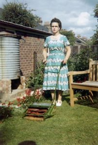 Smartly-dressed middle aged woman standing proudly (if slightly stifly) in a suburban garden with her lawnmower.