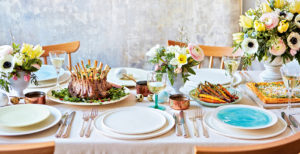 Table laid for an Easter meal with a crown of lamb in the centre with vases of flowers.